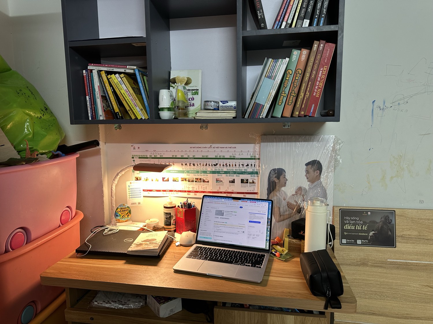 Góc bàn làm việc buổi sáng của Khuất Anh Dũng — đèn bàn, laptop, kệ sách và ảnh gia đình