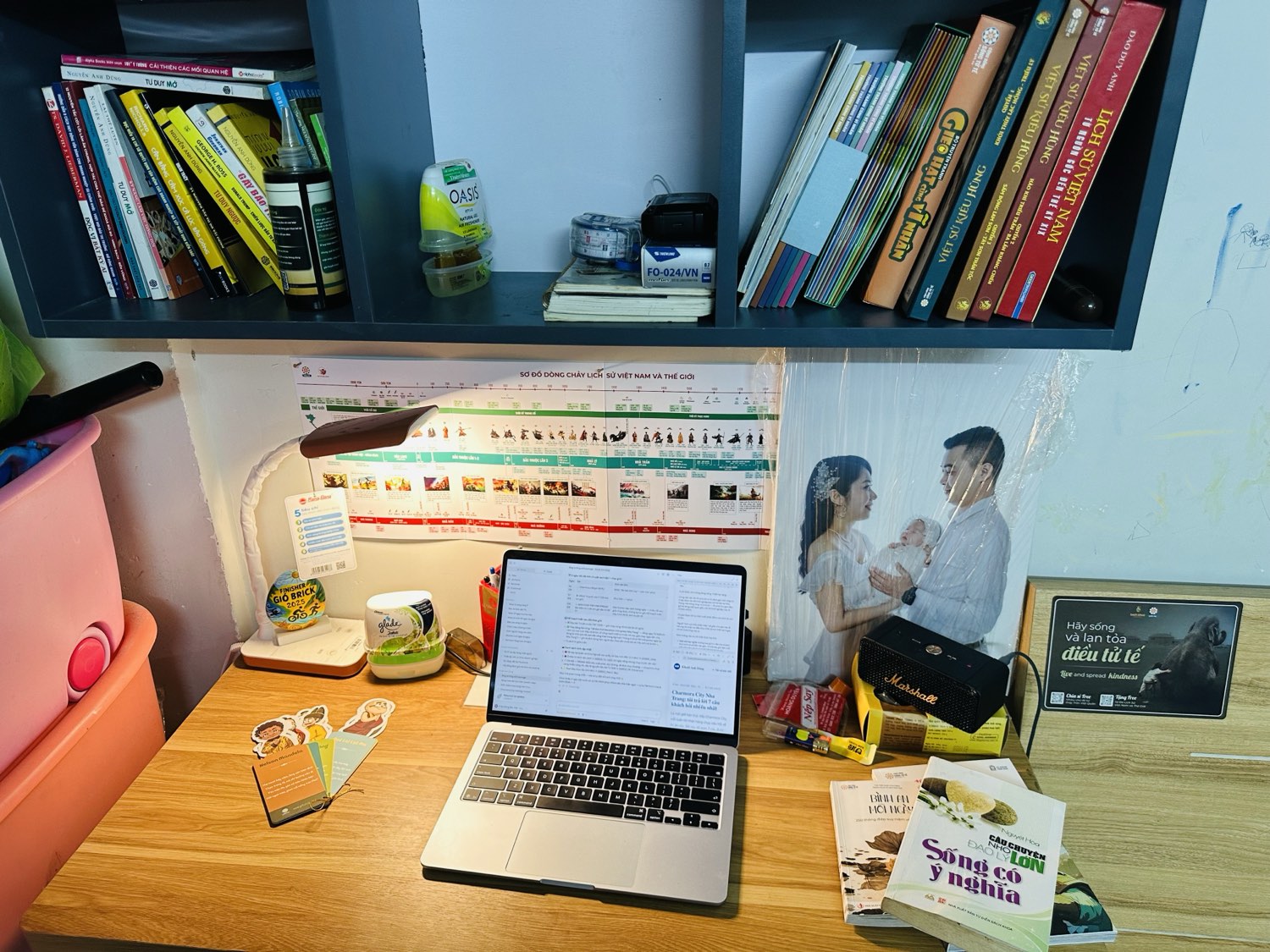 Kệ sách trên bàn làm việc của Khuất Anh Dũng — sách người lớn xen sách thiếu nhi, sơ đồ lịch sử Việt Nam dán tường